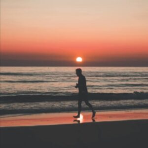 Person jogging on beach at sunset; silhouette against orange sky